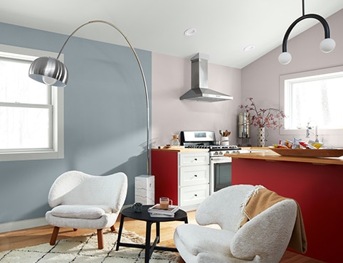 Open living room and kitchen with light blue and lavender-gray walls, a white ceiling, a set of modern white armchairs, and cabinets painted red and white.